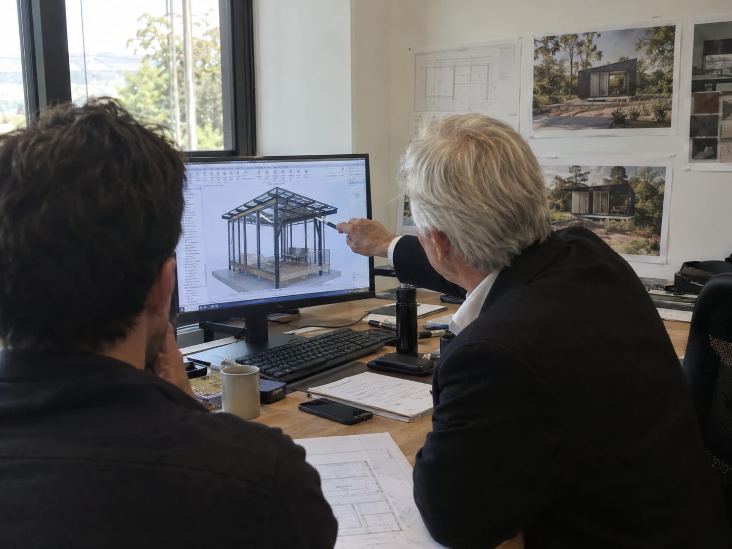 Dos personas de OVO HOMES revisando el modelo 3D de una cabaña en pantalla, con renders exteriores y planos impresos en la pared detrás